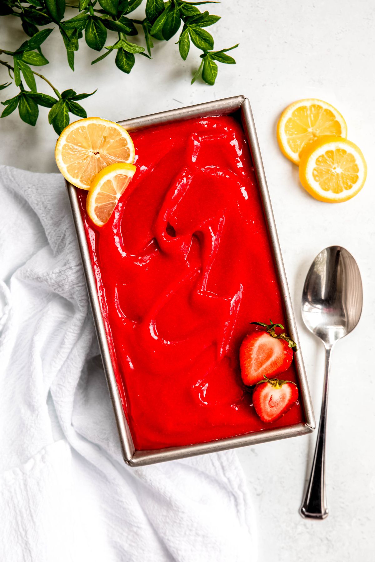 silver bread tin filled with blended strawberry sorbet topped witha. few slices of lemon and a halved strawberry on a white table with more lemon slices and a silver spoon.