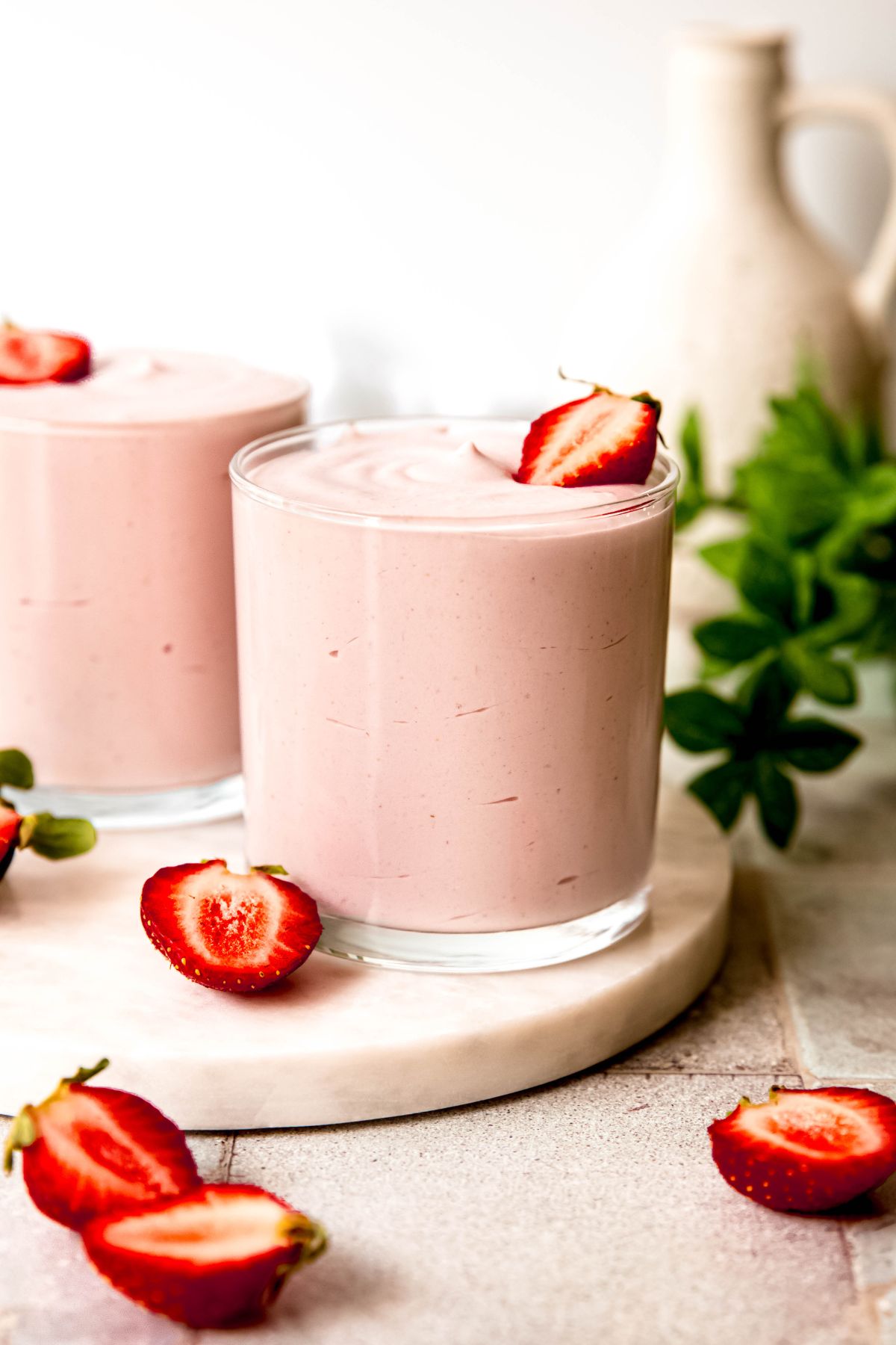 side on view of two glasses of strawberry protein fluff on a white table with halved strawberries and a green houseplant.